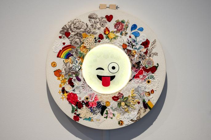 an image of Kenzie Housego's Emoji Bot, which features an emoji winking with its tongue hanging out, lit by LED lights. The emoji is surrounded by embroidered  flowers and other smaller emojis