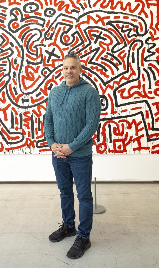 A person smiles while standing in front of a Keith Haring painting with black and red lines on a white background.