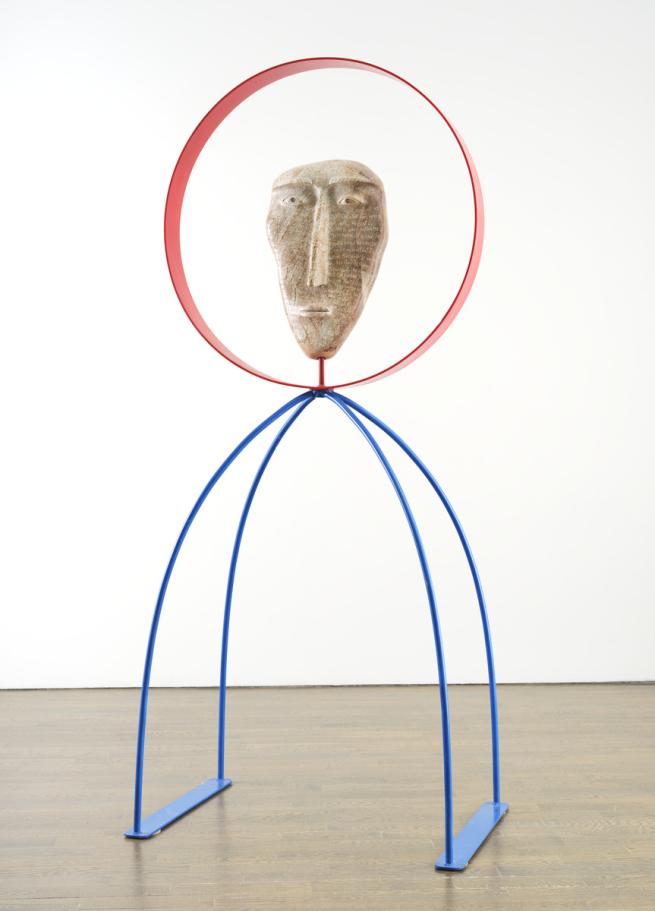 An image of David Ruben Piqtoukun's sculpture People of the Midnight Sun. It features a stone carved face with words carved into it, framed by a red metal circle. The statue stands on two metal blue legs that arch outwards.