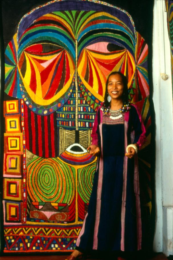 An image of the artist Pacita Abad with her trapunto Bacongo I. She is wearing a dress from Palestine, a necklace from Afghanistan, and tin earrings from Kenya. Bacongo I is a multi-colored mask that
