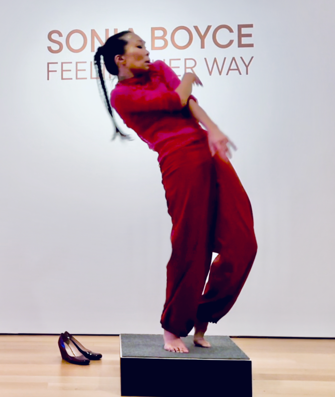 Andrea Naan dancing in front of the Title wall for the exhibition Sonia Boyce: Feeling Her Way. She is on a black platform, and leaning towards the left.