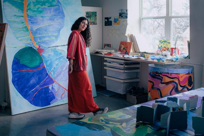 An image of Maru Aponte standing in front of her one of her paintings. She is wearing a red striped top and a solid red skirt and is smiling at the camera