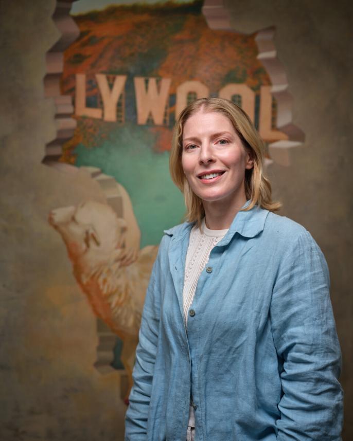 A woman with short blonde hair faces the camera with a smile on her face. She wears a blue button down shirt with a white T-shirt underneath. A painting is in the background.
