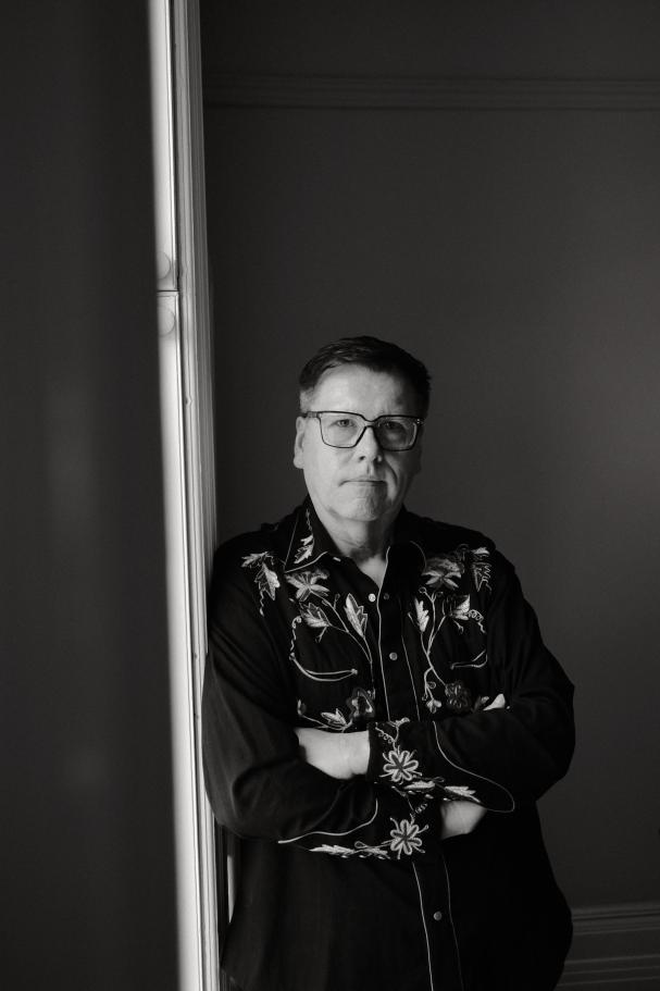 An black-and-white photograph of multidisciplinary artist Barry Ace. He is facing the camera with his arms folded. H
