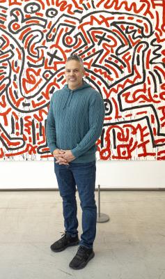 A person smiles while standing in front of a Keith Haring painting with black and red lines on a white background.
