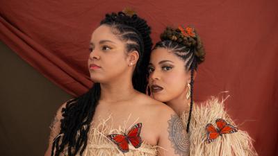 An image of OKAN in front of a dusty orange background with butterflies on their straw outfits