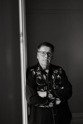 An black-and-white photograph of multidisciplinary artist Barry Ace. He is facing the camera with his arms folded. H