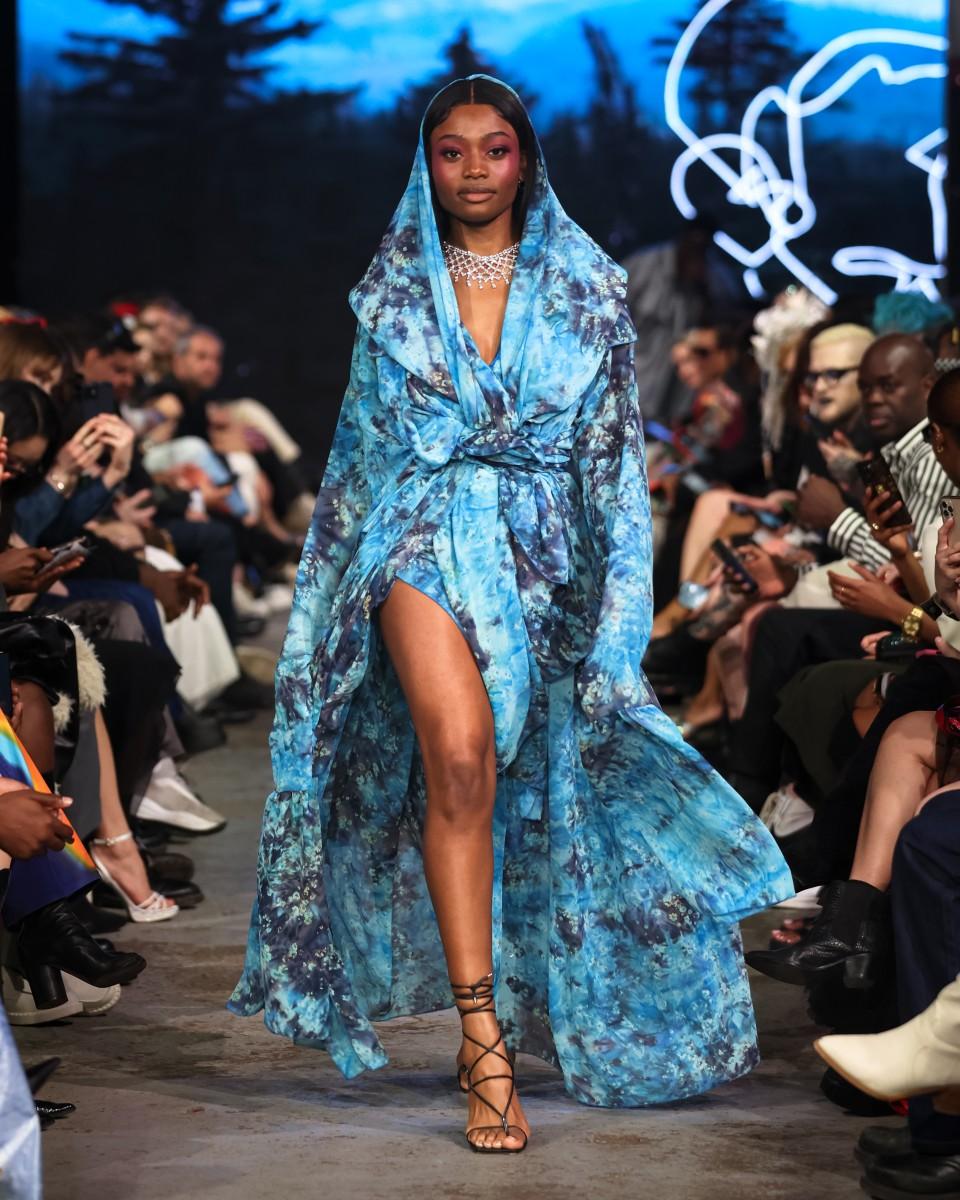 a model walking down a runway in a blue dress designed by Mic. Carter for their fashion line L’Uomo Strano