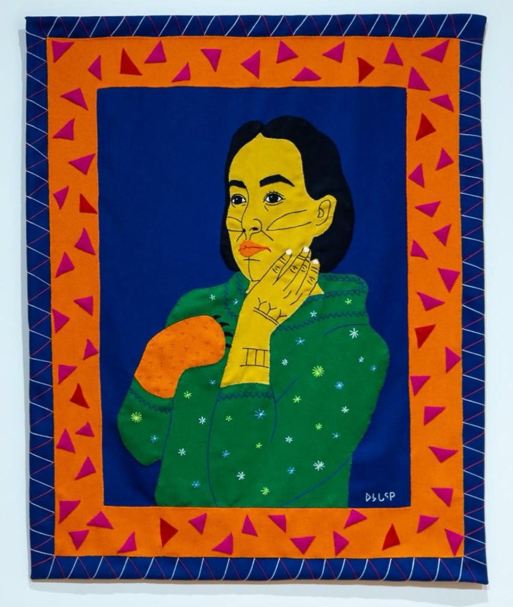 An image of Artist Gayle Uyagaqi Kabloona's piece Tiriganiaq, which features a woman with kakiniit on her face and left hand, which rests on her face. Her other hand is a fox paw and she wears a green polka dot sweater against a dark blue background. The image is framed first in an orange frame with pink triangles, then a dark blue frame with light blue and pink geometric triangles