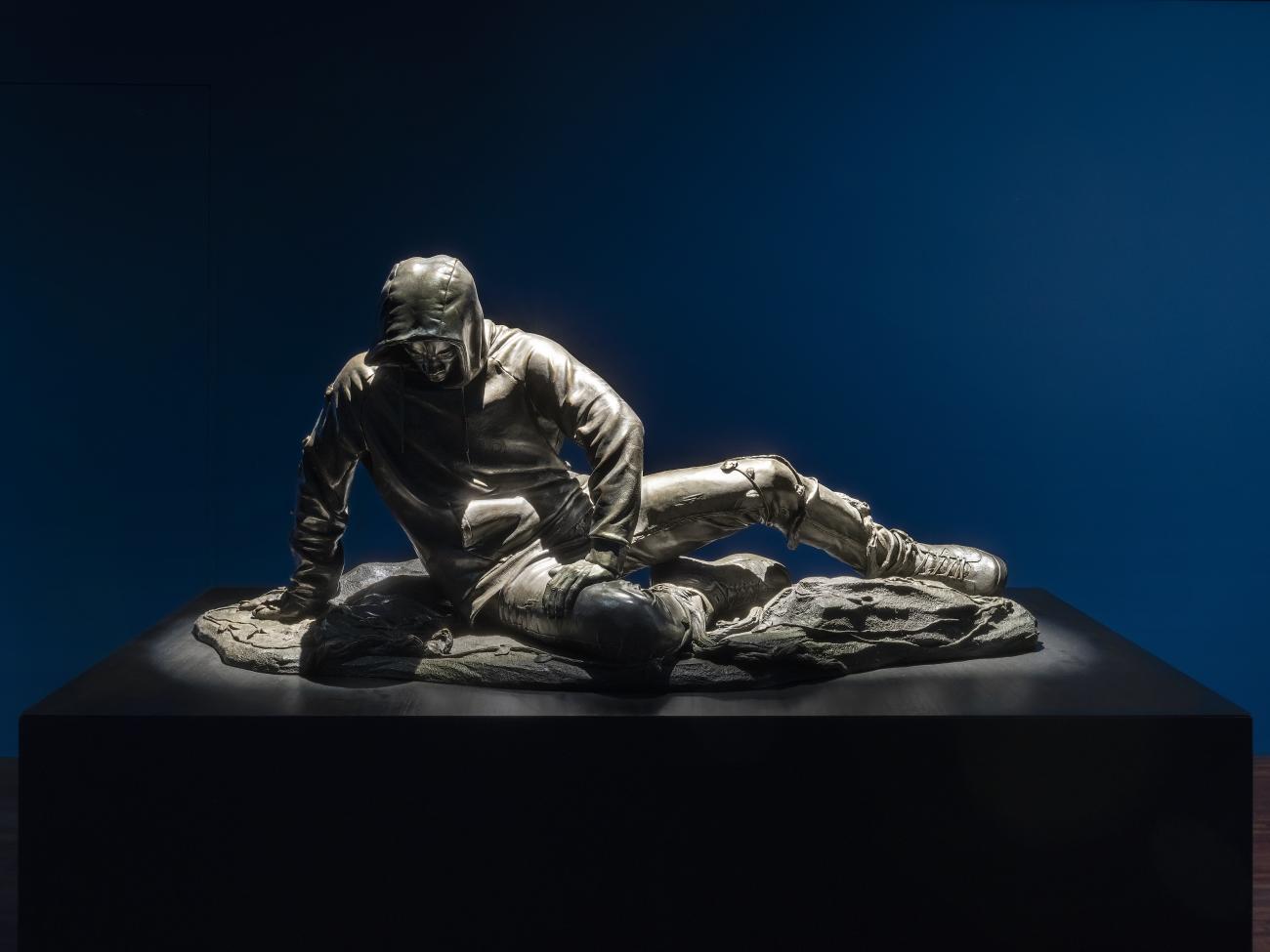 An image of Kehinde Wiley's sculpture Dying Gaul, after a Roman Sculpture of the 1st Century, which features a young black man in a hoodie with his hood up, propping himself up on the ground with one arm and his legs laid horizontally. His face is turned downwards.