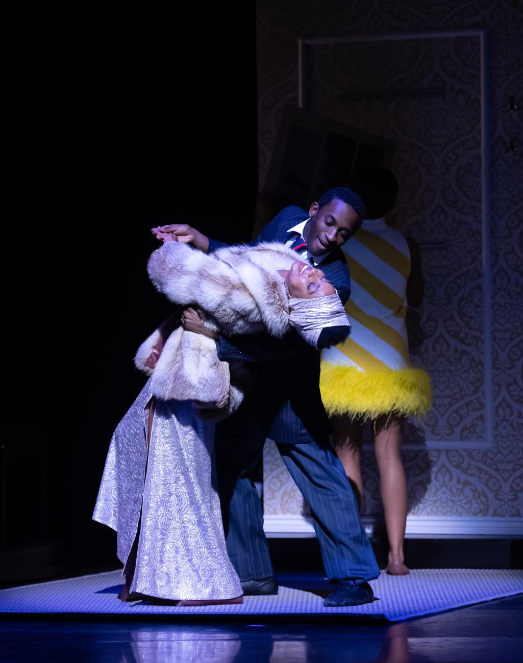 Two dancers from Ballet Black dancing together on stage. One dancer in a suit dips the other dancer in an ornate dress and fur coat