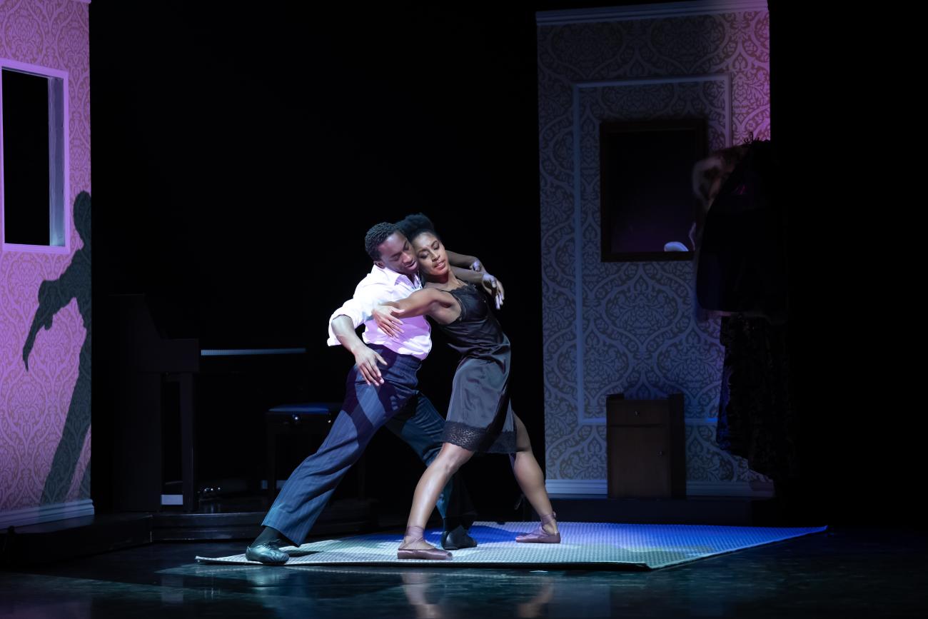 Two dancers from Ballet Black dancing together on stage. One dancer in a black dress leans her head on the shoulder of a dancer in a white dress shirt behind her