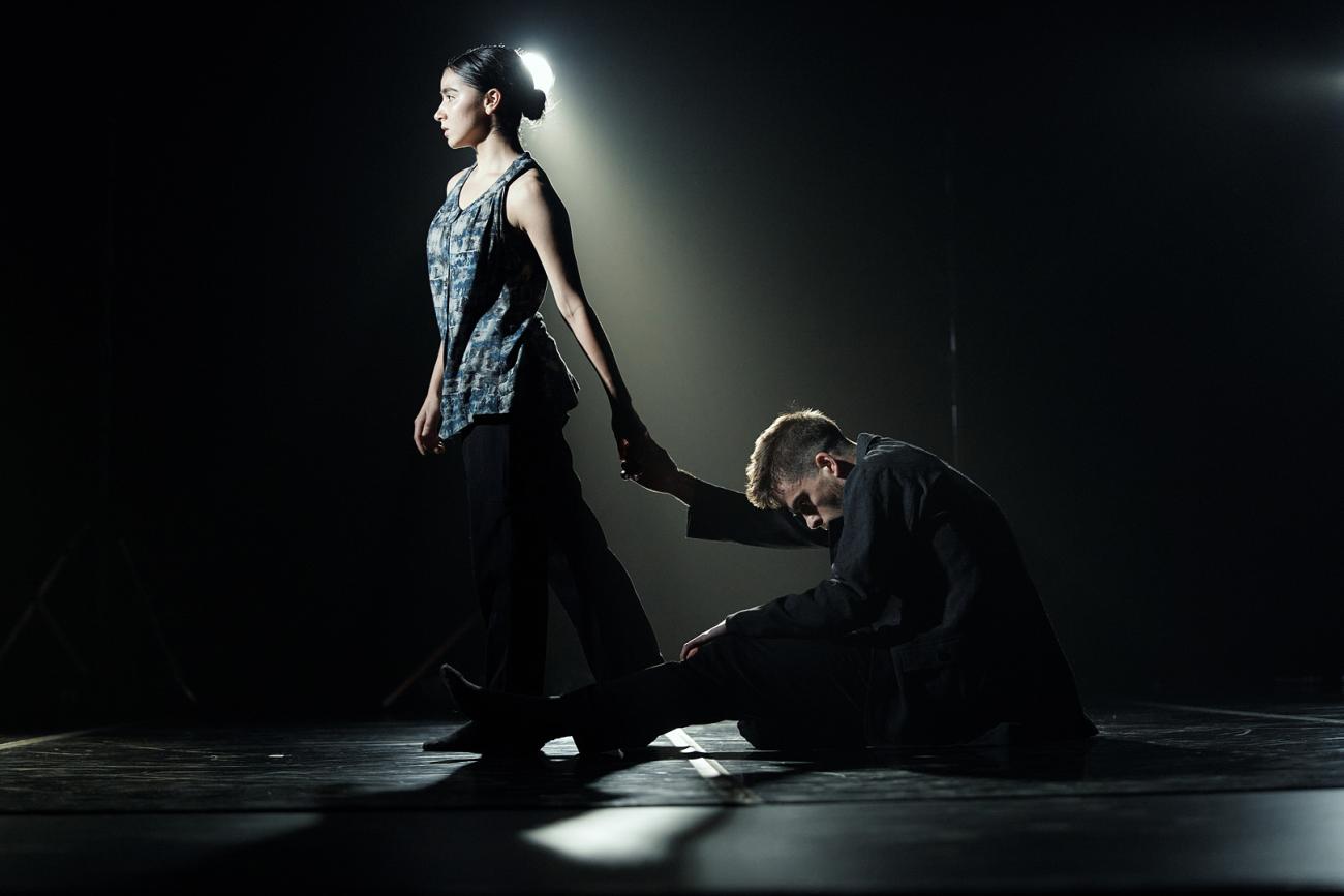 Two dancers on stage, one dancer sits on the floor with his head looking downwards. The other dancer holds his outstretched hand, walking forward