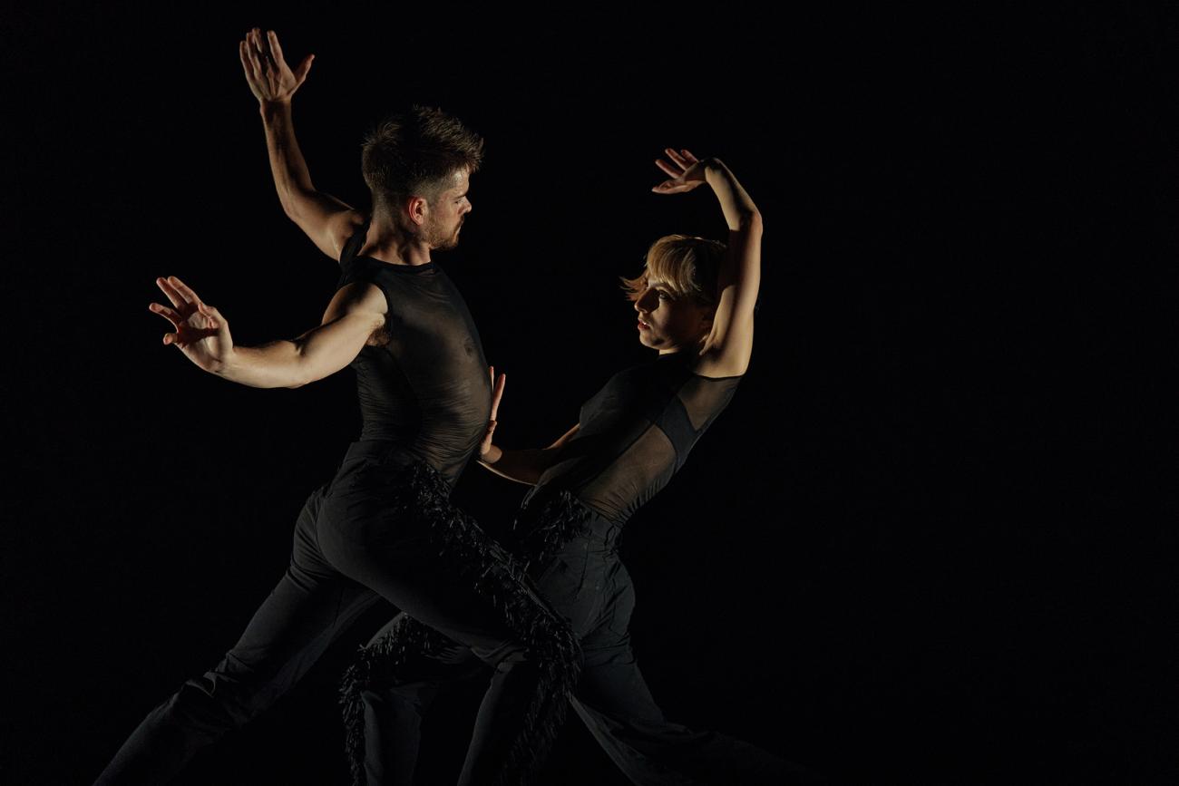 Two dancers on stage in all black facing each other, their hands are up and one dancer leans backwards while the other dancer leans towards them