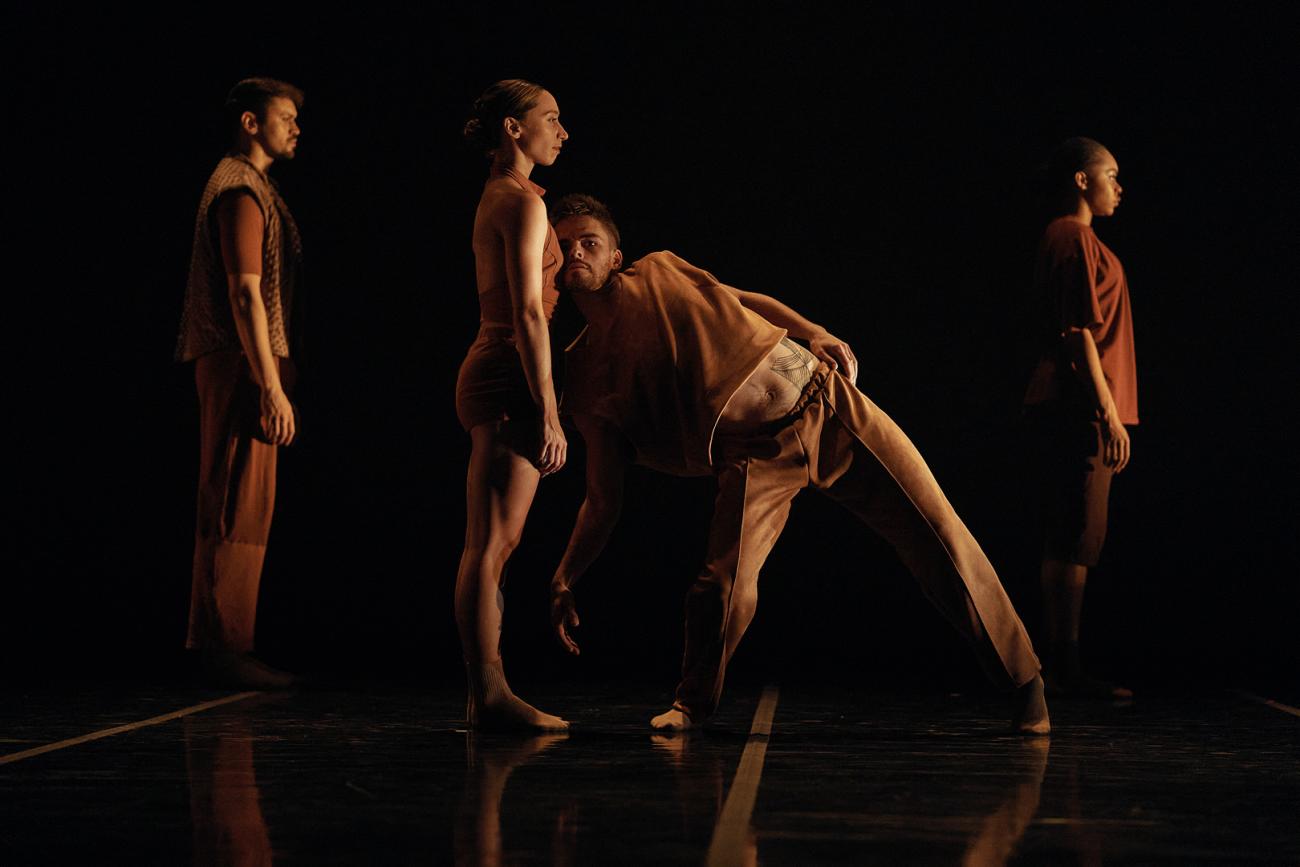 An image of dancers on stage, facing the right. One dancer faces the audience, leaning their head on the torso of the other dancer