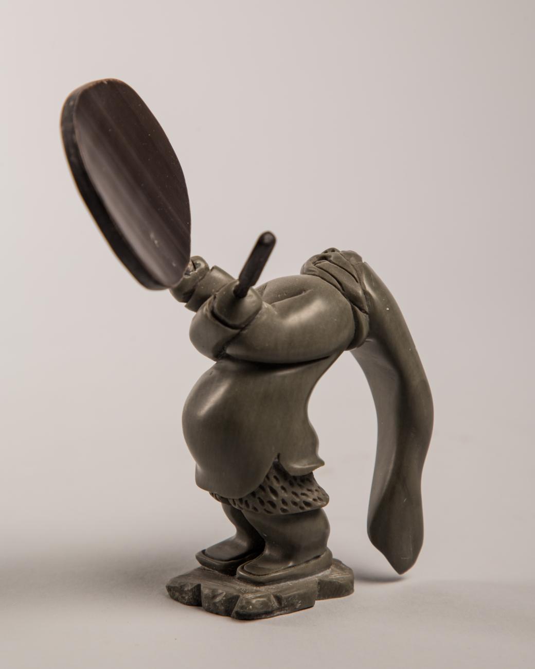 A sculpture of a shaman drum dancer. They hold a drum in one hand, and a stick in the other.