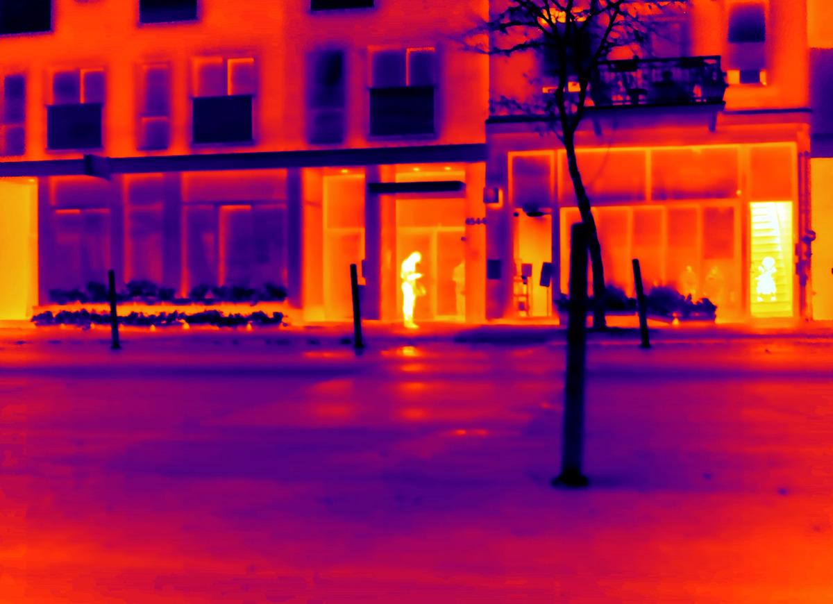 An image of Emmanuelle Léonard's Le livreur et sa cliente, 22:53/ Delivery person and Client, 10:52 pm. The photograph is taken with a thermal camera. A delivery person stands on the street looking at their phone, their silhouette bright yellow. A person waits for their delivery in the building on the staircase, which glows a bright yellow