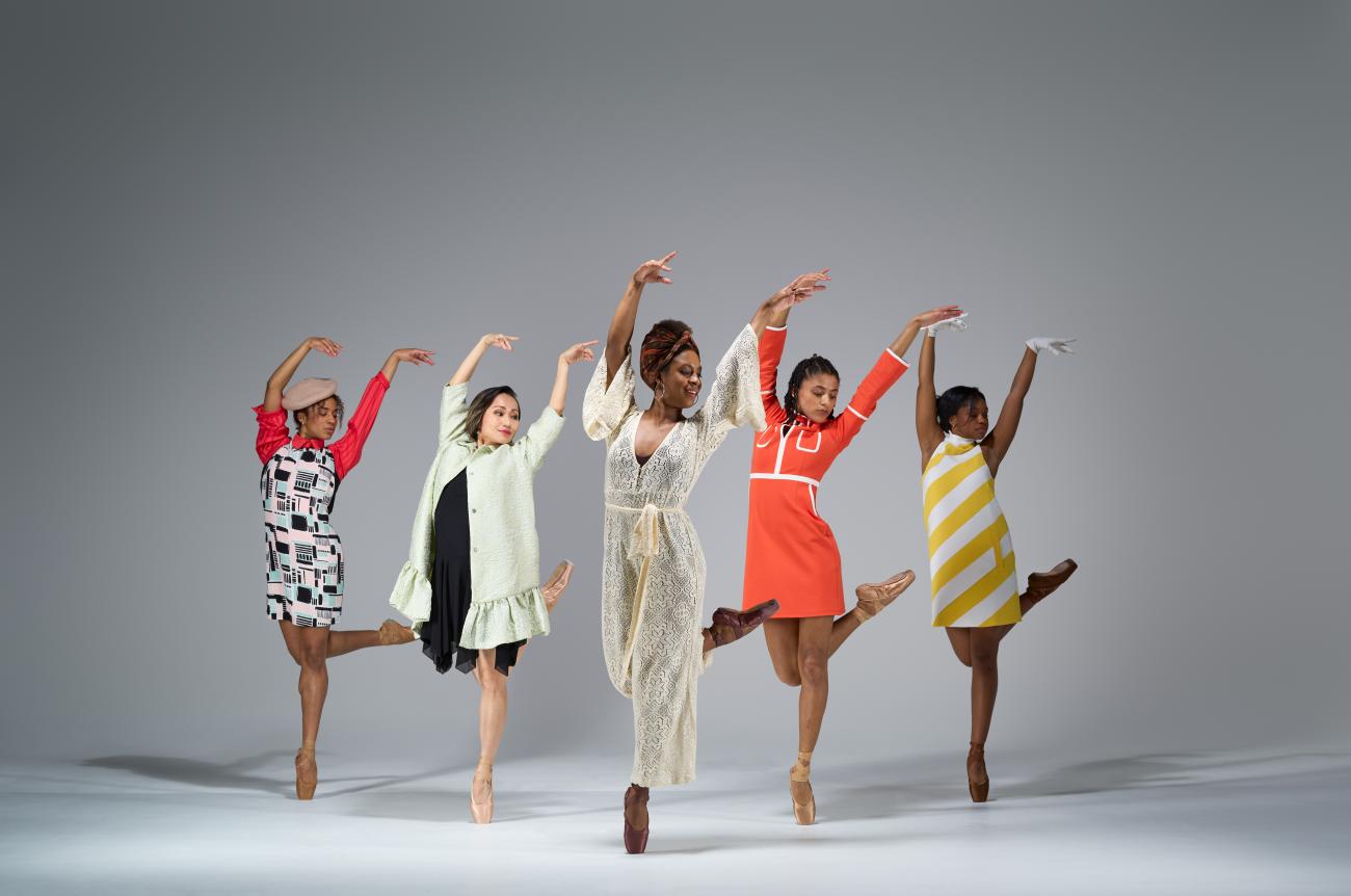 five dancers in a triangle formation posing in pointe shoes with their right foot bent upwards behind them, their arms up in the air with their hands bent to their right. Their heads are slight turned to the right. They are dressed in vintage looking outfits.