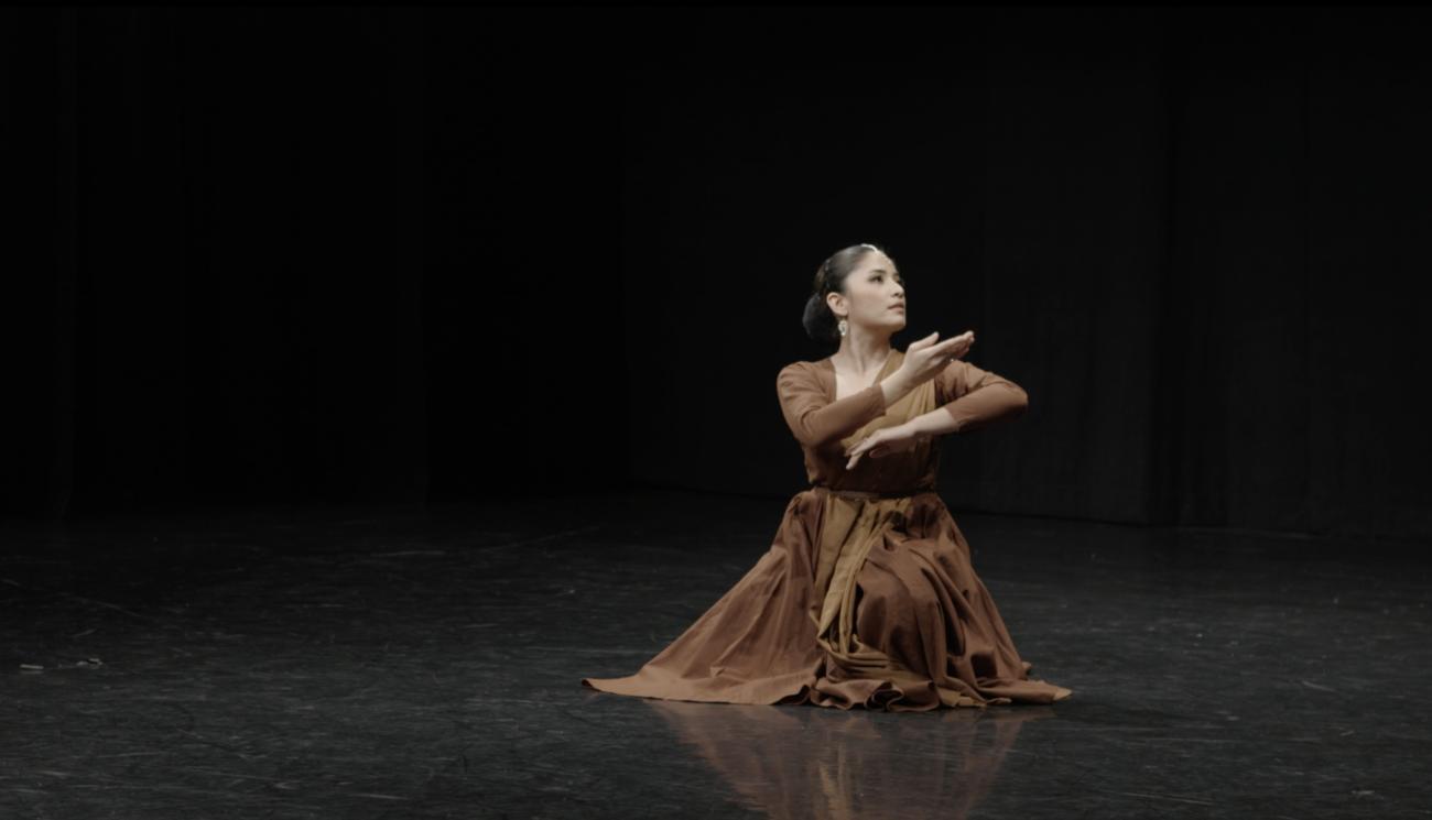 Dancer Fiana Kawane dancing on stage. She sits on the floor looking to their left, their hands are folded and posing near their chest.
