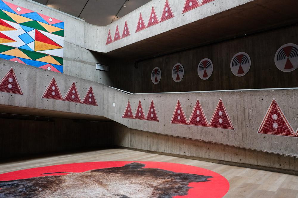 An installation image of Niitoyis, featuring a large vinyl of a buffalo hide on the floor, and images of bead work and parfleche on the walls