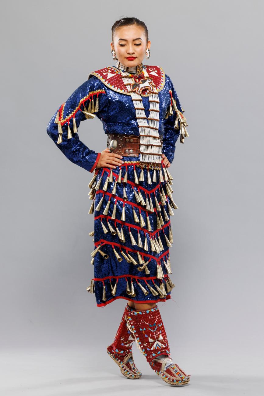 An image of a Jingle Dress Dancer Posing. Her dress is a shiny blue with red highlights. She is wearing red boots and her gaze is pointed downwards