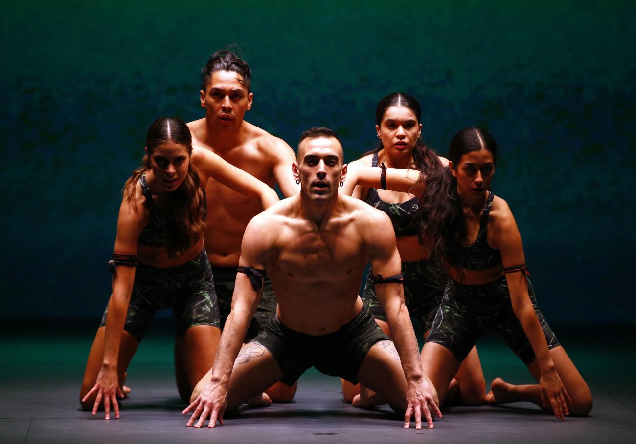 An image of performers from Red Sky Performance. 5 dancers are on their knees in a diamond formation, the other dancers touching the back of the dancer in the front