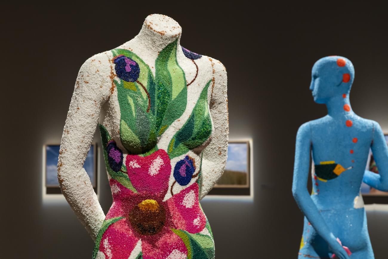 an image of two beaded mannequins, one with white with a large pink flower, leaves, and purple berries. The second has its back to the camera, its blue and has orange circles running down its head
