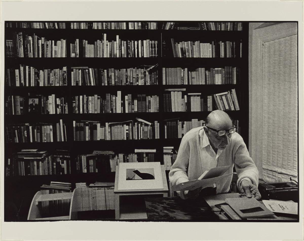 Stravinsky seated at a desk and surrounded by books. He wears a button-up shirt and glasses.