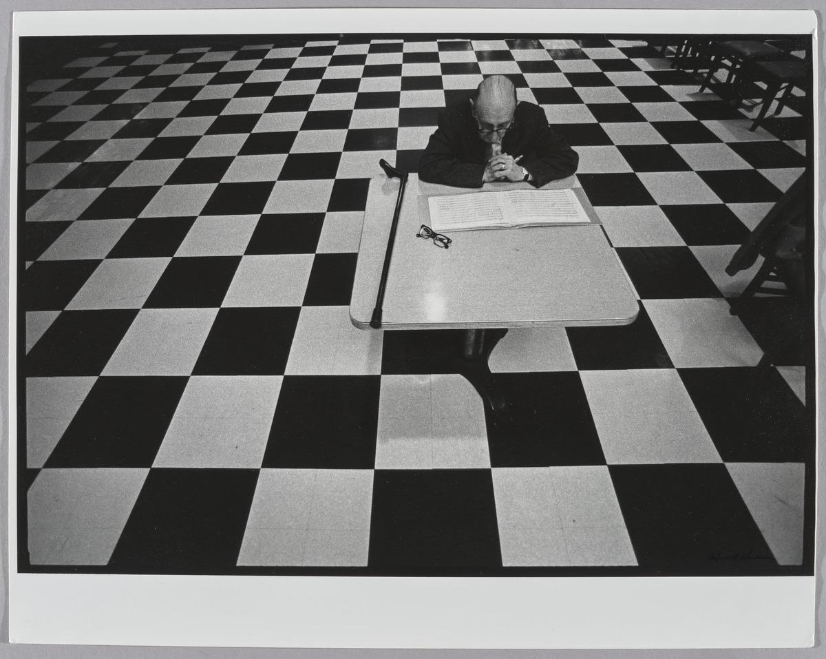 Stravinsky sitting at table, reading some papers. The floor is checkered.