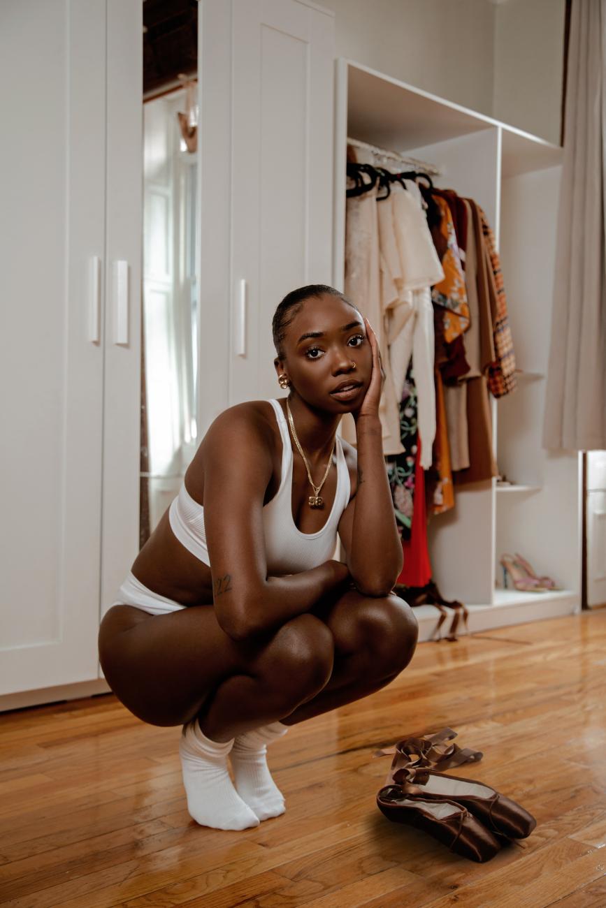A photograph of dancer Jordan Peterson crouching and posing in front of her closet and beside her pointe shoes
