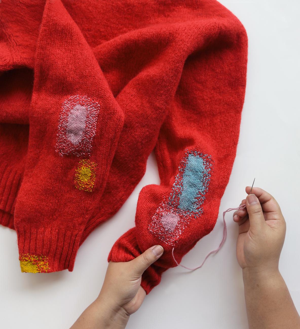 A photograph of a red sweater mended with pink, yellow, and blue stitching on the elbows