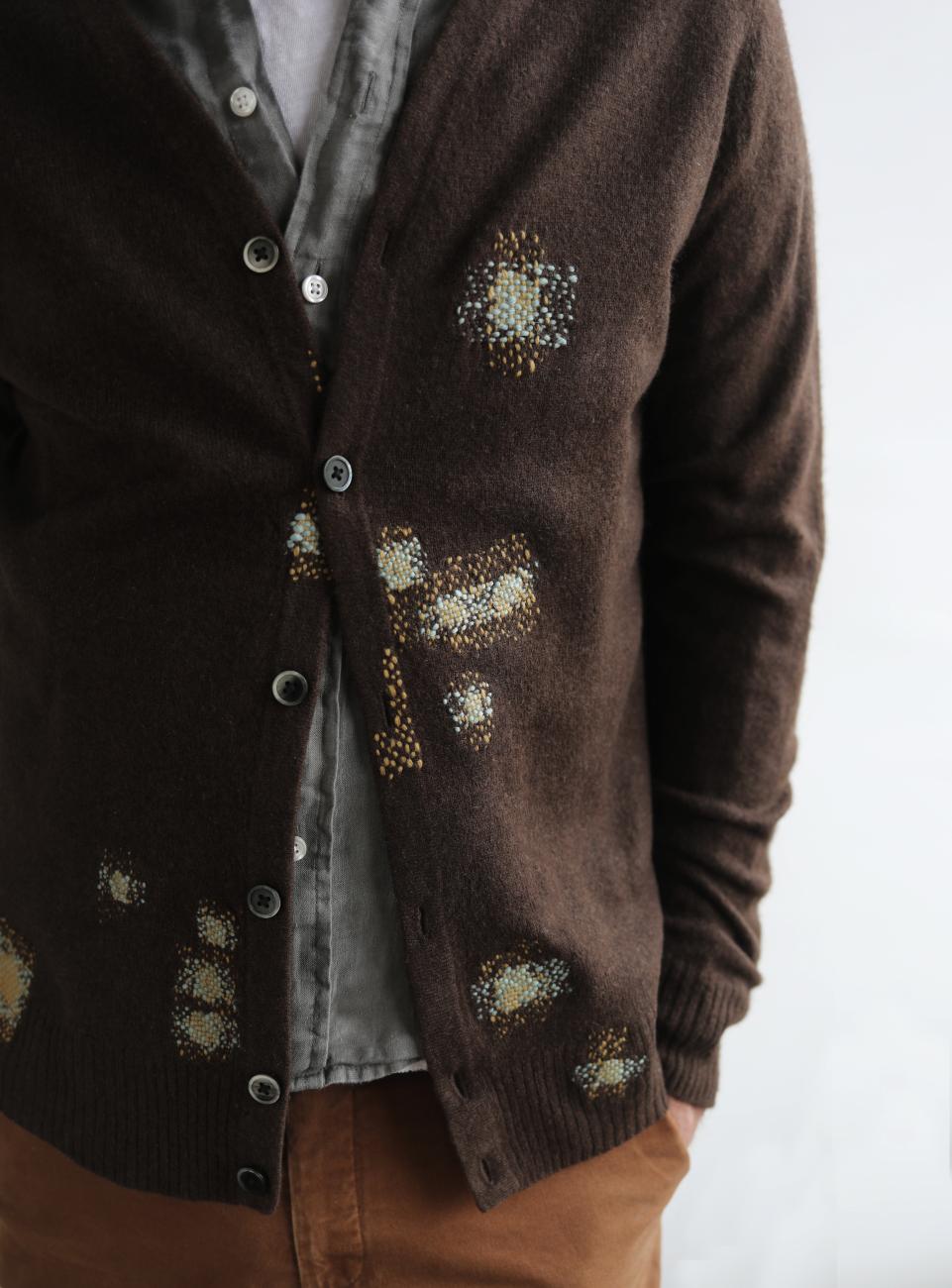 An photograph of someone wearing a brown sweater mended in various places with a light green and yellow thread