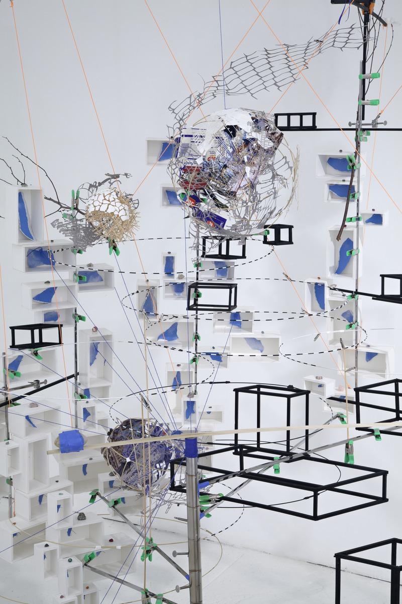 An image of Sarah Sze's sculpture Disappearing Act (2012). It is a close up of a section of the sculpture featuring orbs made with eggshells, toothpicks, and paper amongst various white cuboids filled with torn blue paper