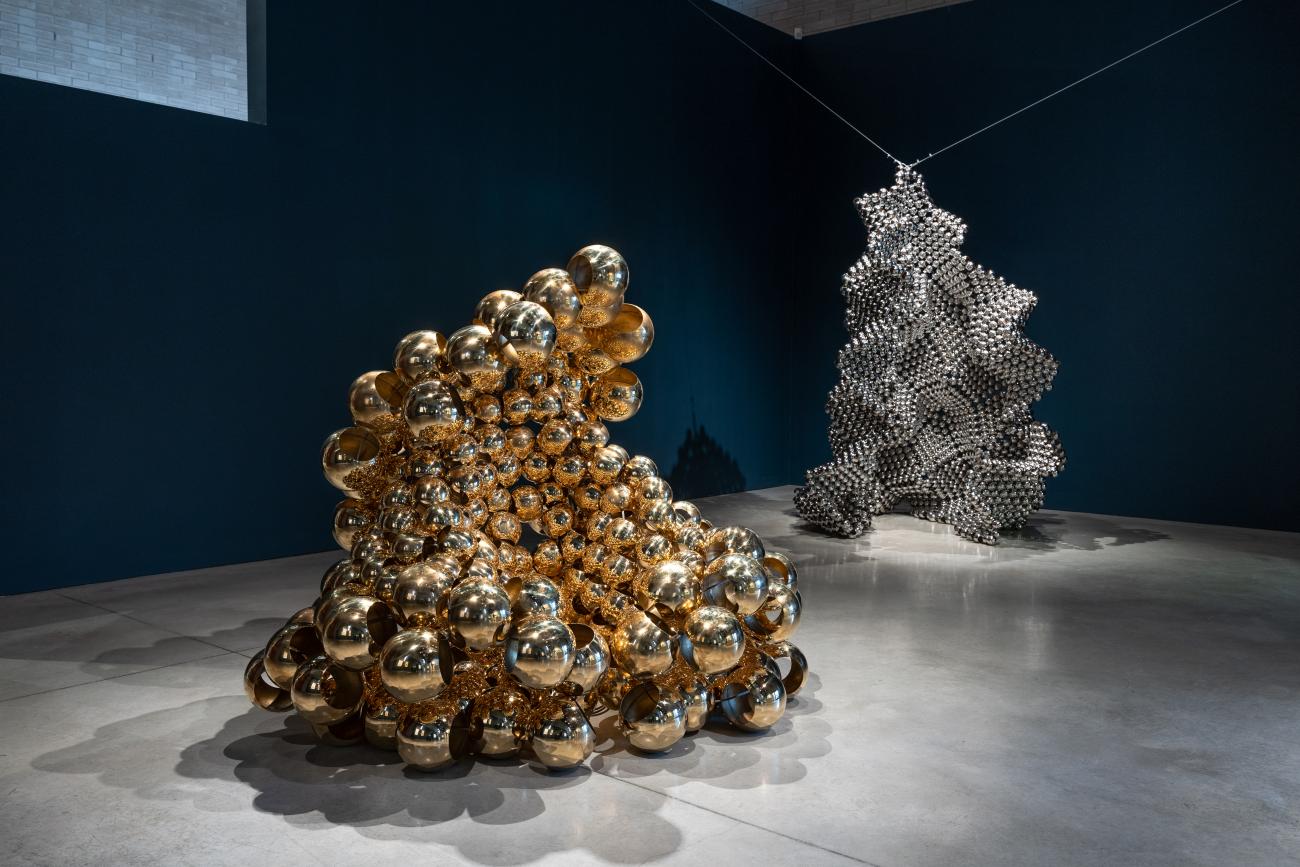 an installation image of two sculptures in Sharl G. Smith's exhibition Beyond the bead. The sculpture in the foreground is made with gold metallic beads while the sculpture in the background is created with silver metal beads and is suspended from the ceiling
