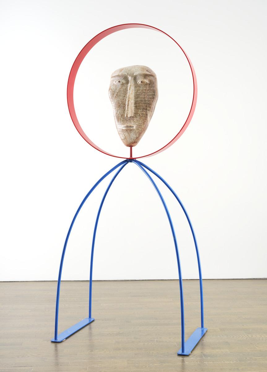 An image of David Ruben Piqtoukun's sculpture People of the Midnight Sun. It features a stone carved face with words carved into it, framed by a red metal circle. The statue stands on two metal blue legs that arch outwards.
