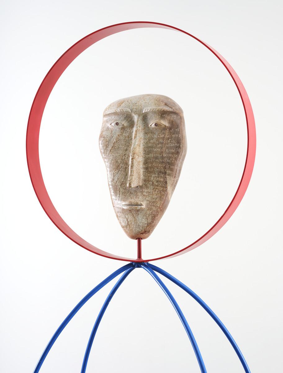 A detail shot of David Ruben Piqtoukun's sculpture People of the Midnight Sun. This photograph is a close up of the stone carved face, which has words carved all over it. The face is framed in a red metal hoop.