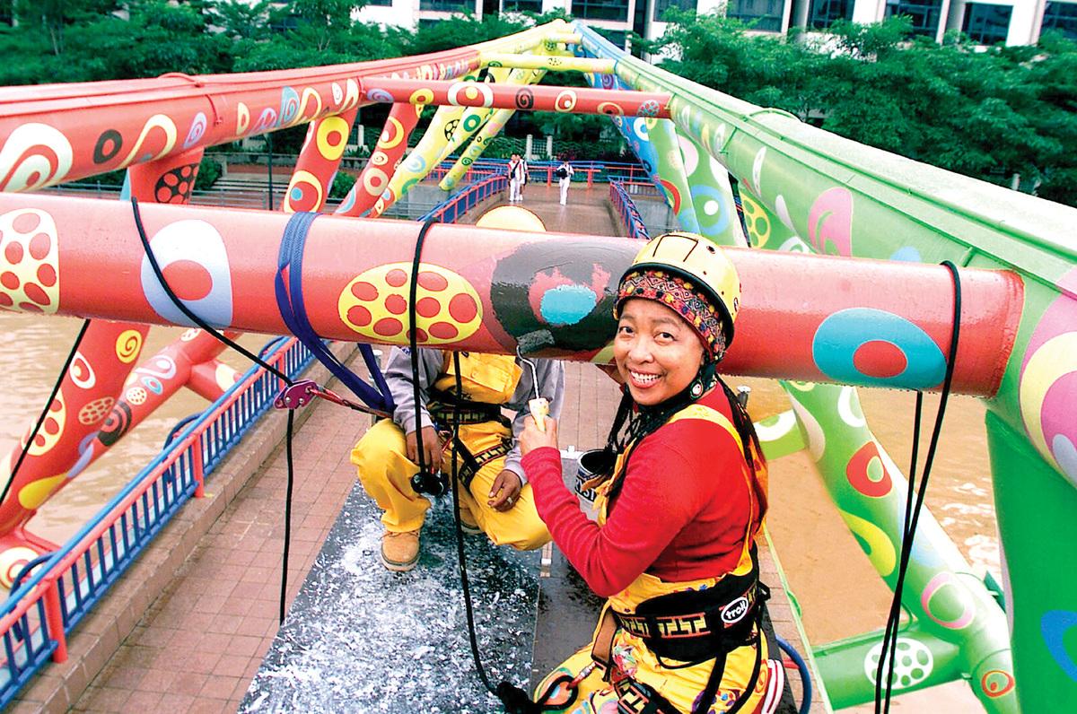 An image of Pacita Abad painting the Alkaff Bridge in Singapore. She is in a harness and helmet, turning around to face the camera mid-paint
