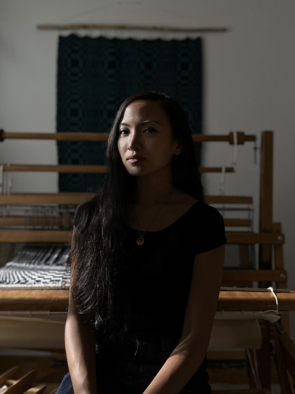 An photograph of Carol Ann Apilado in her studio. She sits posed in front of her loom and one of her weaved works hanging on the wall