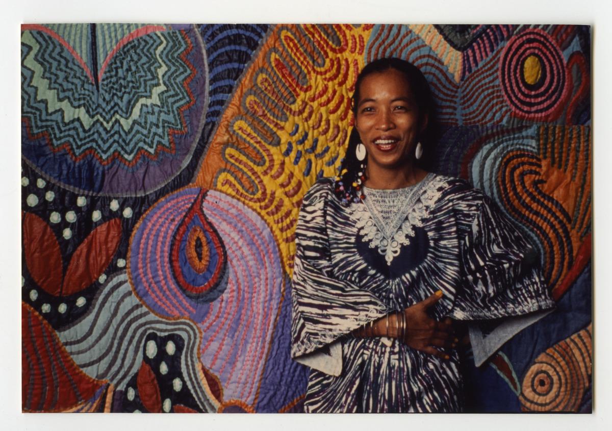 An image of Pacita Abad with her trapunto painting Ati-Atihan. She is wearing clothing and accessories collected during her travels.