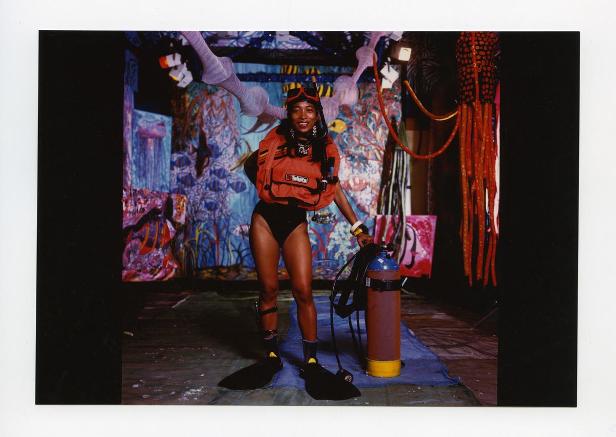 A photograph of artist Pacita Abad at her exhibition opening for Assaulting the Deep Sea. She is wearing a bathing suit and her scuba diving gear, including a life jacket, slippers, googles, and snorkels. She poses beside her oxygen tank.