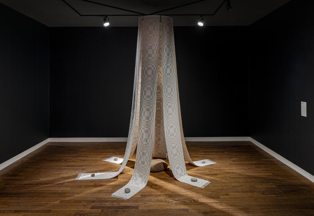 An image of Carol Ann Apilado's work Channel (2023). It features strips of woven fabric hanging from the ceiling like a canopy. As these strips meet the floor, they are weighed down by rocks.