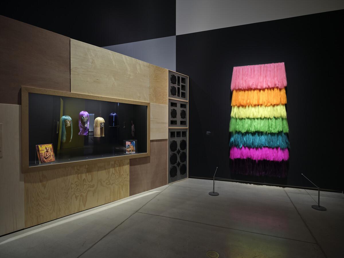 An installation shot of Lil' Kim's wigs, re-created by Dionne Alexander. To the right of the wigs is auntie fawn on tha 6, which features neon pink, orange, yellow, green and blue neon hair