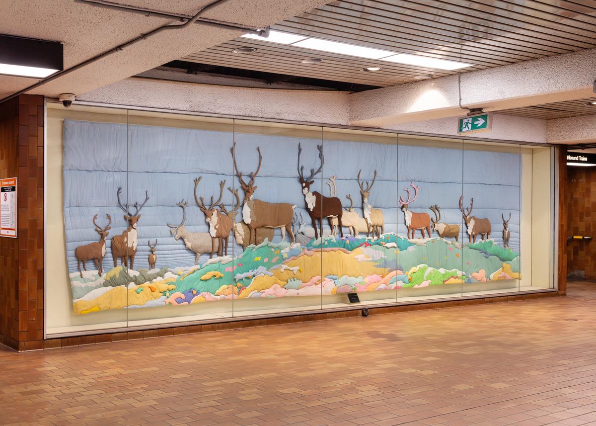 An image of Joyce Wieland's quilt Barren Ground Caribou installed in Spadina Station. The quilt features numerous caribou in a multicoloured field