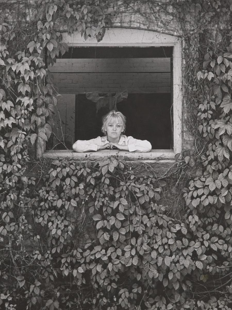 A black-and-white photograph of Joyce Wieland posing in the window of an ivy-covered building
