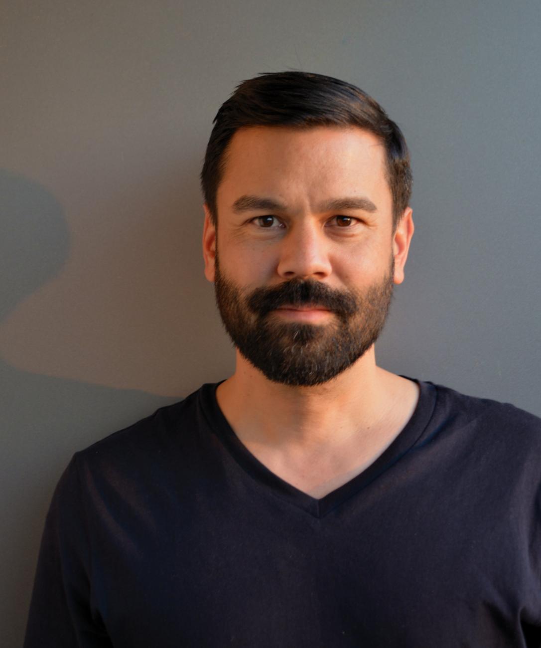 A photo of the artist Jon Sasaki. He is closed mouth smiling at the camera and is wearing a black v-neck