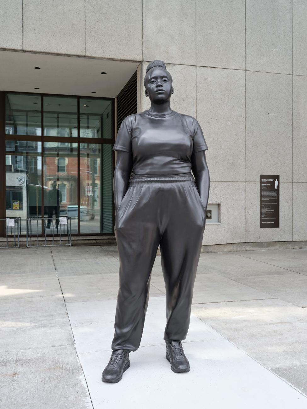 A hyperrealistic sculpture of a young Black woman dressed casually with her hands in her pockets, looking out into the distance.