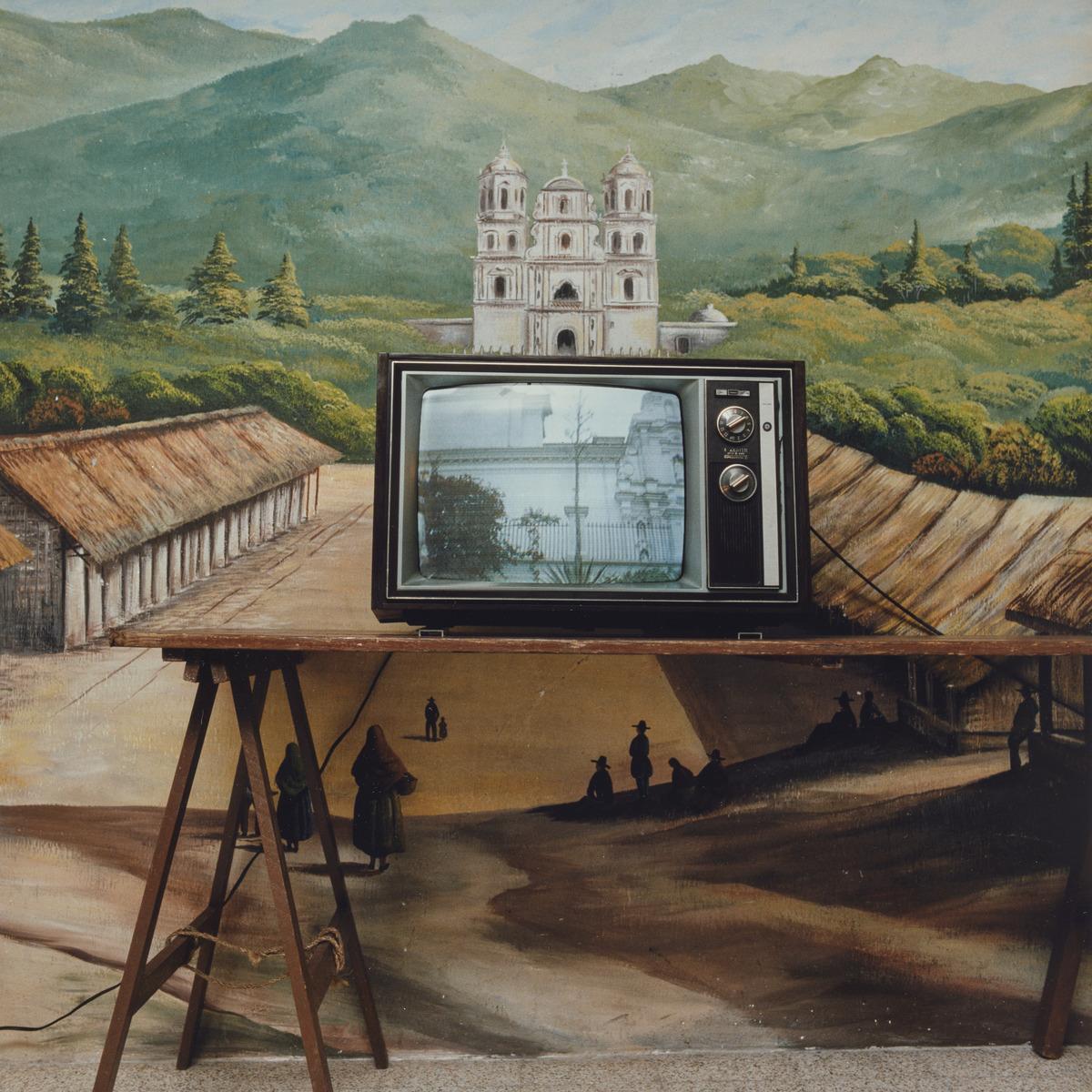 An image of Rafael's photograph Press Lounge, Esquipulas, Guatemala. It features a black & white television that is on and placed on a table in front of a mural.