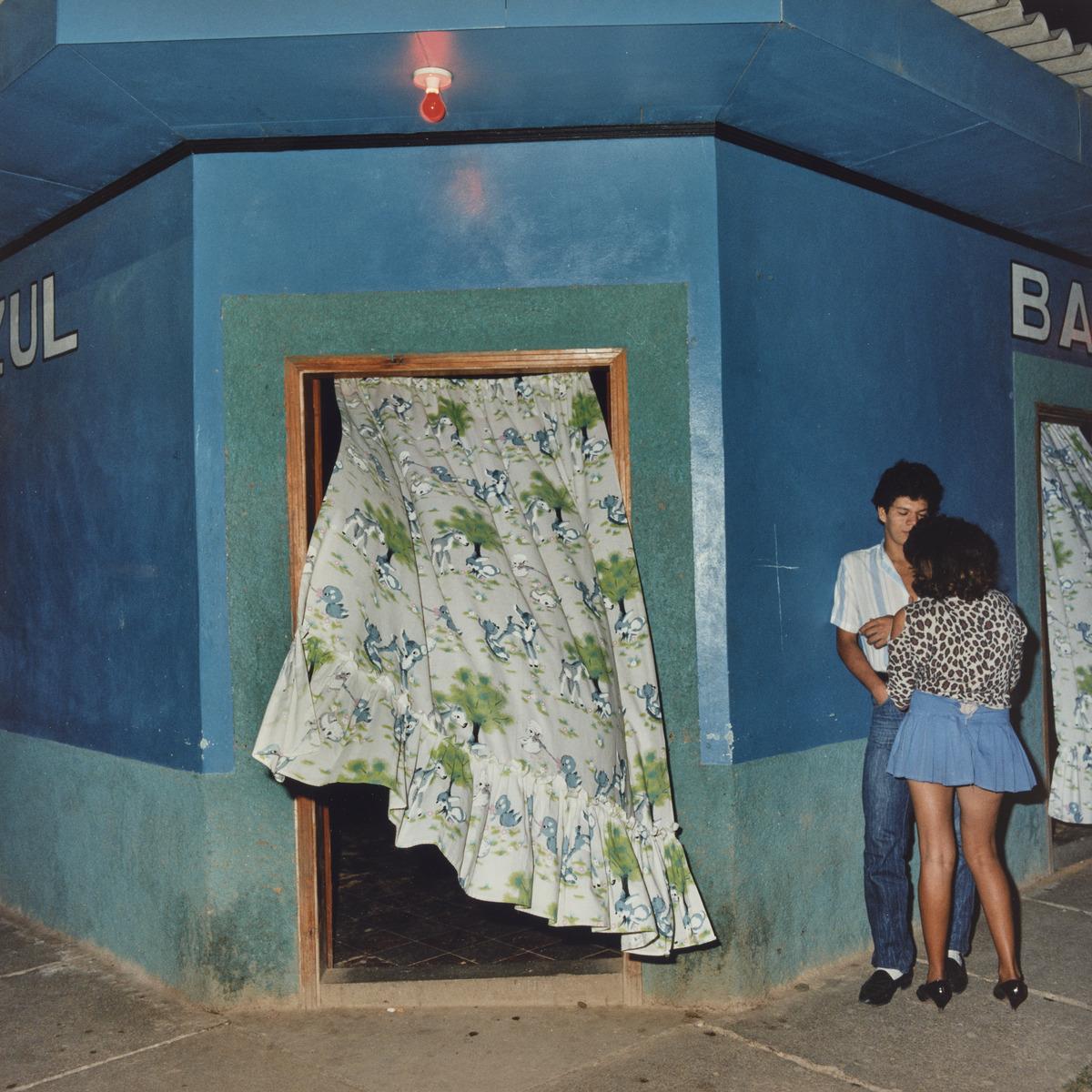 An image of Rafael Goldchain's photograph Nocturnal Encounter, Comayagüa, Honduras. It shows a couple standing facing each other outside a blue building with a curtain billowing out of the doorway.