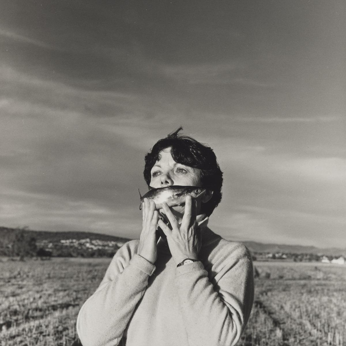 An image of Graciela Iturbide's photo Autorretrato en el campo, Pachuca, México (Self-Portrait in the field, Pachuca, Mexico). It is a self-portrait of Iturbide with a fish over her mouth