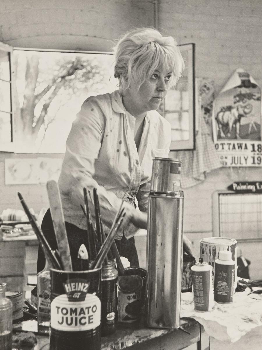 A black and white photograph of Joyce Wieland in her studio. She is looking away from the camera with her hands busy